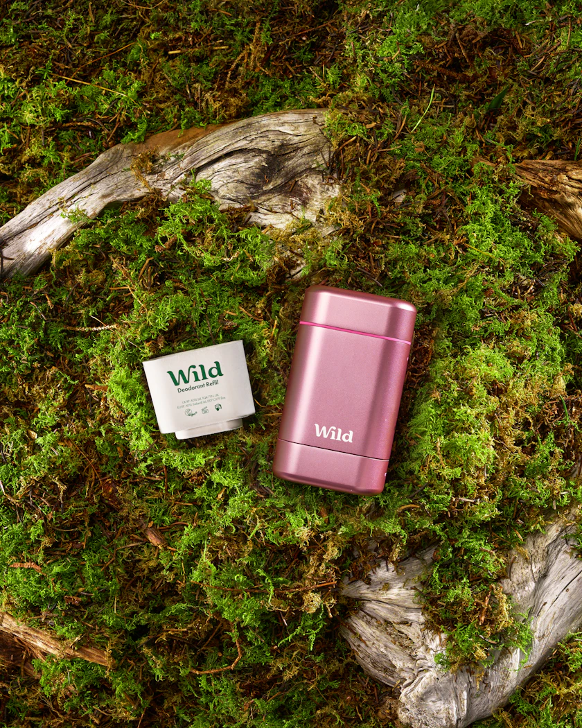 Two Wild deodorants, one pink stick and one white refill pouch, on mossy ground with driftwood.