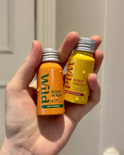 A hand holds two small bottles of Wild Cosmetics body wash. One bottle is orange labeled Juicy Mango, the other is yellow labeled Sweet & Almond.