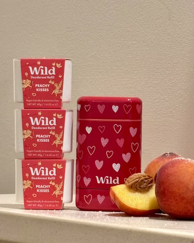 Three pink deodorant refills labeled "Peachy Kisses" and a red deodorant container with white hearts, alongside peaches and pecans.