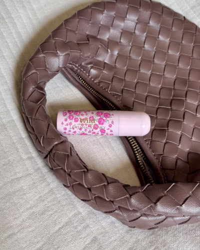 A pink lipstick tube with floral patterns sits on a brown woven purse.